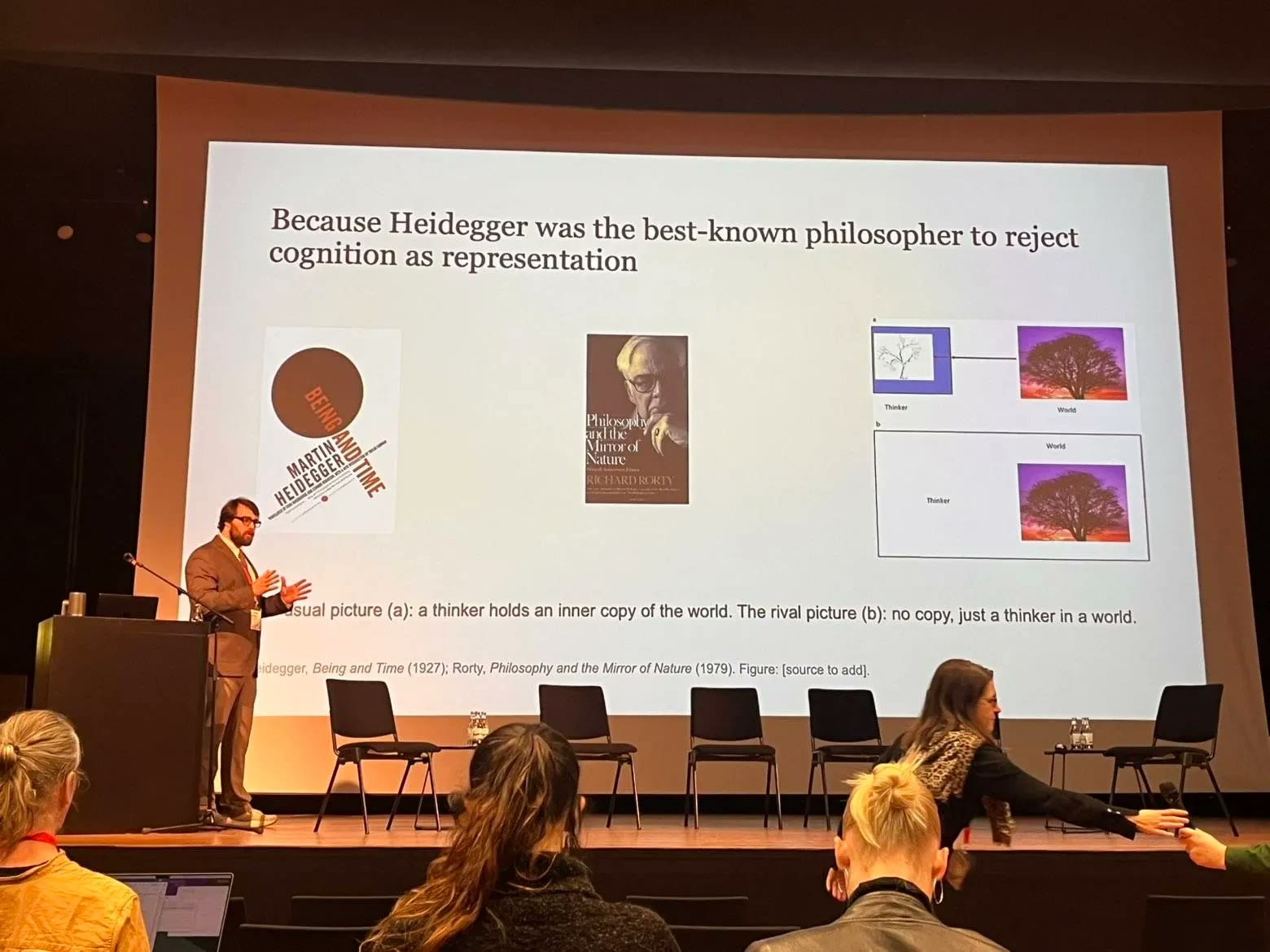 Collin Lucken presenting at SAB 2026 in Berlin, in front of a slide about Heidegger and the rejection of cognition as representation